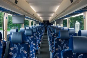 Spacious motorcoach interior with rows of blue patterned seats, overhead lighting, and overhead monitors