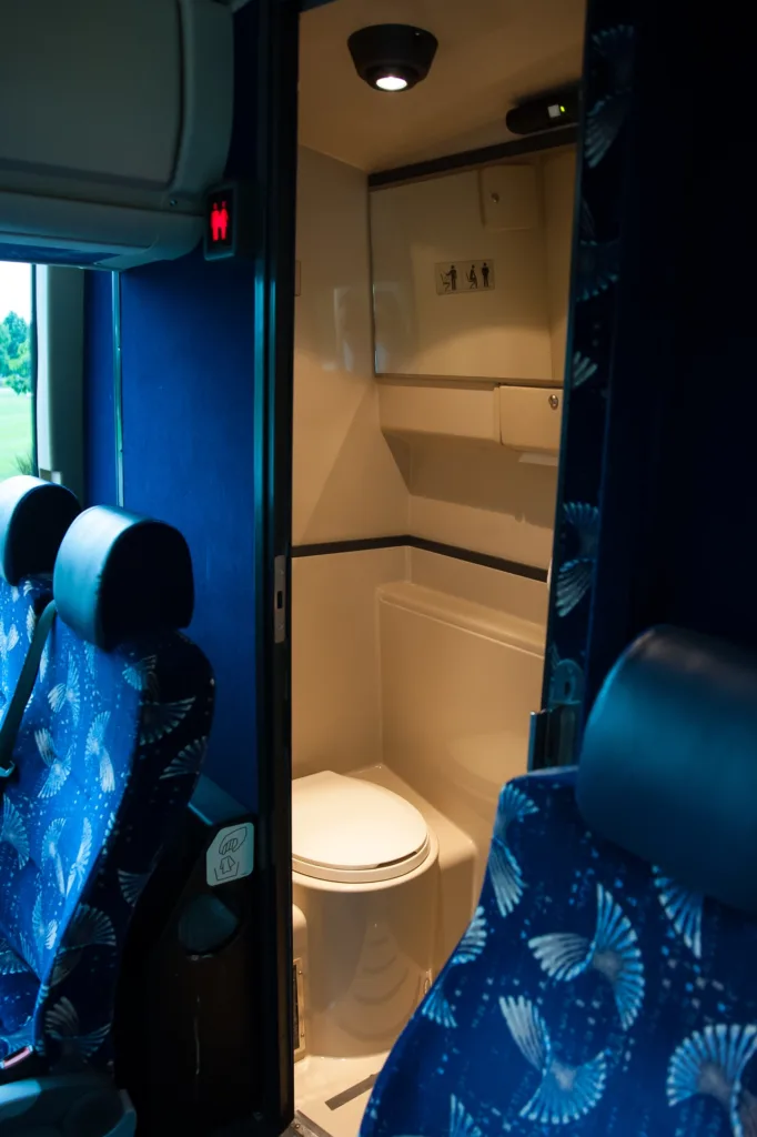 Onboard motorcoach restroom with toilet, overhead light, and occupied indicator sign visible from the cabin